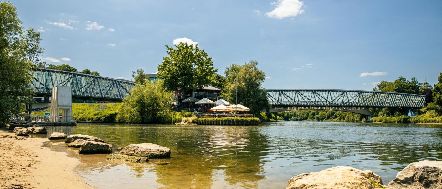 Idyllisches Flussufer in Remseck mit Sandstrand, einer modernen Brücke und einem Café mit Sonnenschirmen unter Bäumen., © Stuttgart-Marketing GmbH, Sarah Schmid Idyllisches Flussufer in Remseck mit Sandstrand, einer modernen Brücke und einem Café mit Sonnenschirmen unter Bäumen., © Stuttgart-Marketing GmbH, Sarah Schmid
