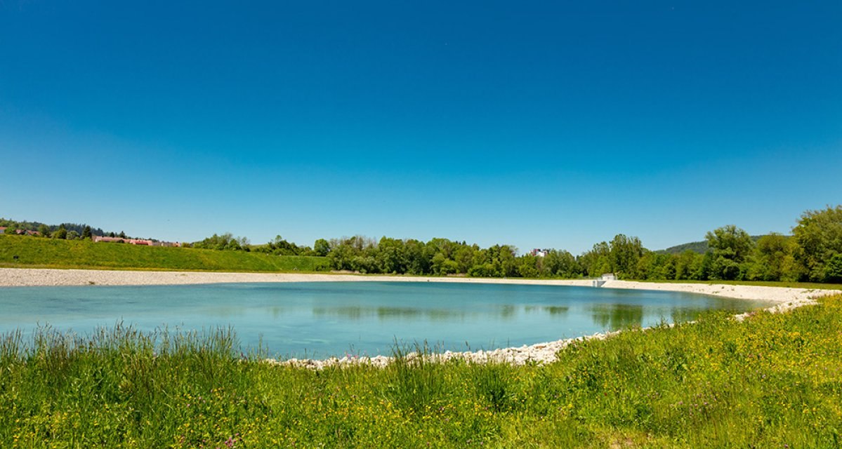 Das Bild zeigt das Unterbecken des Naturstromspeichers in Gaildorf, umgeben von gr&uuml;ner Vegetation und einem klaren blauen Himmel.