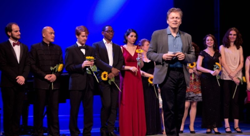 Künstler auf einer Bühne bei Rossini in Wildbad. Einige halten Sonnenblumen, während ein Mann in der Mitte spricht. Der Hintergrund ist blau beleuchtet., © Touristik Bad Wildbad GmbH Künstler auf einer Bühne bei Rossini in Wildbad. Einige halten Sonnenblumen, während ein Mann in der Mitte spricht. Der Hintergrund ist blau beleuchtet., © Touristik Bad Wildbad GmbH