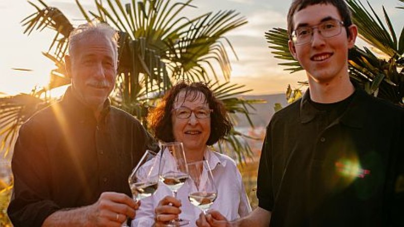 Drei Personen sto&szlig;en mit Weingl&auml;sern an, umgeben von Palmen im Sonnenuntergang., &copy; Weingut Bayer