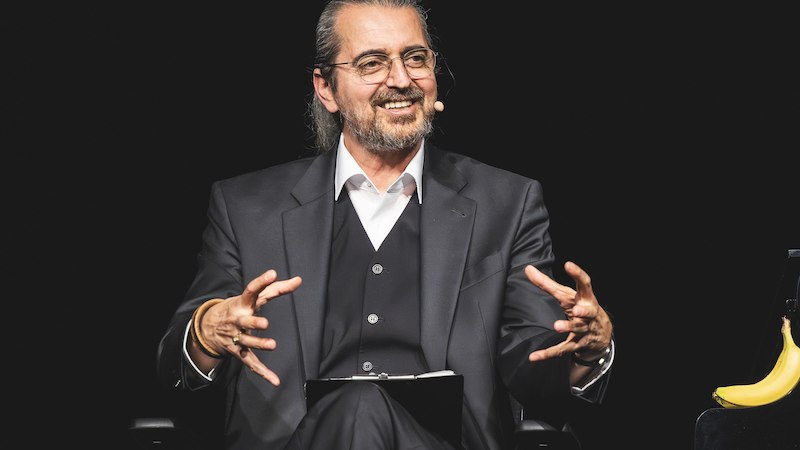 Ein Mann in einem dunklen Anzug sitzt auf einer Bühne, spricht in ein Mikrofon und gestikuliert. Neben ihm liegt eine Banane., © Scala Kultur Live gGmbH