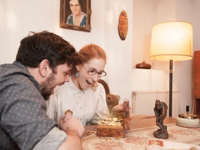 Ein Mann und eine Frau freuen sich &uuml;ber eine ge&ouml;ffnete Schatztruhe voller M&uuml;nzen in einem gem&uuml;tlichen Raum., &copy; ExitGames Stuttgart