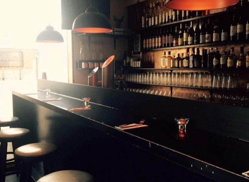 Gem&uuml;tliche Bar mit dunkler Theke, Barhockern und Regalen voller Flaschen. Warmes Licht schafft eine einladende Atmosph&auml;re., &copy; Emilio's
