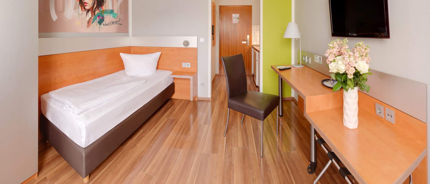 Modernes Einzelzimmer mit Bett, Schreibtisch, Stuhl und Wandkunst. Helle Holzböden, Flachbildfernseher und Blumen auf dem Tisch., © attimo Hotel Stuttgart