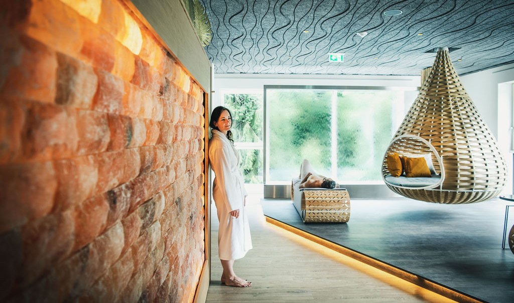Moderner Saunaraum mit Salzsteinwand, H&auml;ngesessel und Liege. Eine Person in Bademantel lehnt an der Wand, eine andere liegt entspannt auf der Liege., &copy; LOCHER Fotodesign&Manufaktur