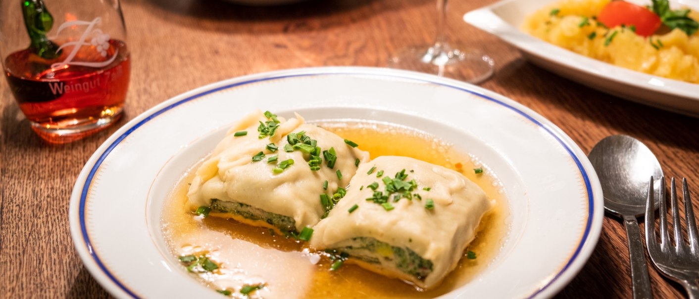 Zwei Maultaschen in Brühe auf einem Teller, garniert mit Schnittlauch. Daneben ein Glas Rotwein und Besteck auf einem Holztisch., © Weingut Zaiß