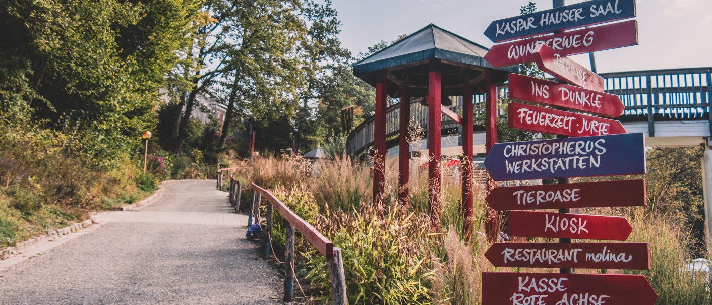 Ein Wegweiser mit roten und blauen Schildern zeigt verschiedene Ziele in einem grünen Park an. Ein Weg führt durch die Landschaft., © Stadt Welzheim Ein Wegweiser mit roten und blauen Schildern zeigt verschiedene Ziele in einem grünen Park an. Ein Weg führt durch die Landschaft., © Stadt Welzheim