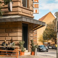 Eine Straßenecke mit einer Metzgerei. Zwei Personen sitzen an einem Tisch vor dem Laden. Ein Auto fährt vorbei, und es gibt eine Ampel., © SMG, Sarah Schmid Eine Straßenecke mit einer Metzgerei. Zwei Personen sitzen an einem Tisch vor dem Laden. Ein Auto fährt vorbei, und es gibt eine Ampel., © SMG, Sarah Schmid
