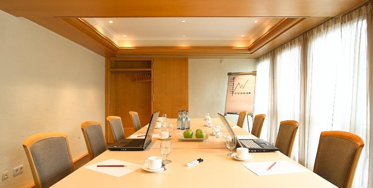 Konferenzraum mit Tisch, St&uuml;hlen, Laptops, Notizbl&ouml;cken und Wasserflaschen. Flipchart mit Diagramm im Hintergrund, helle Vorh&auml;nge an den Fenstern., &copy; Hotel am Park