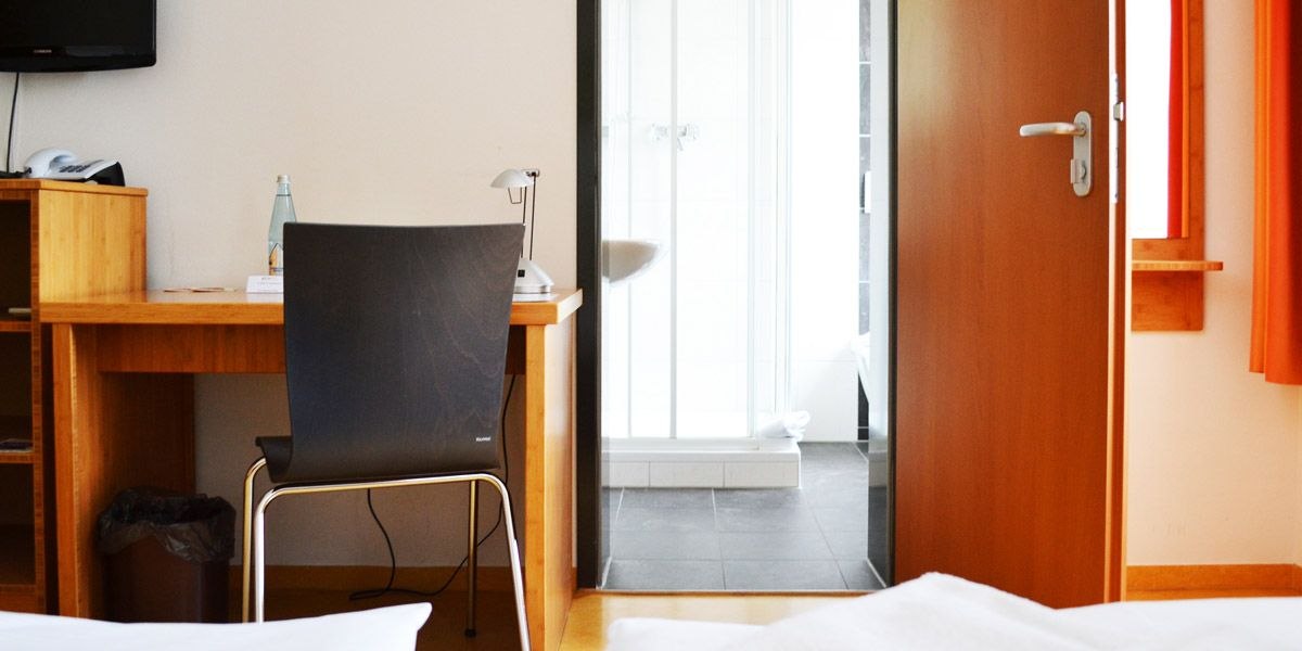 Ein Hotelzimmer mit Schreibtisch, Stuhl und Blick ins Badezimmer. Eine Wasserflasche und Lampe stehen auf dem Tisch., &copy; ECOINN Hotel am Campus