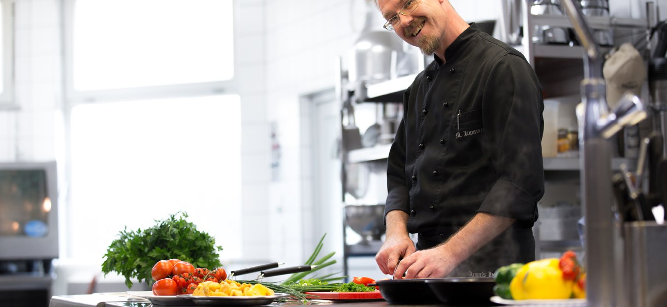 Ein Koch in schwarzer Uniform schneidet Gemüse in einer hellen, professionellen Küche. Frische Zutaten liegen auf der Arbeitsfläche., © ARAMIS Hotel & Freizeit GmbH