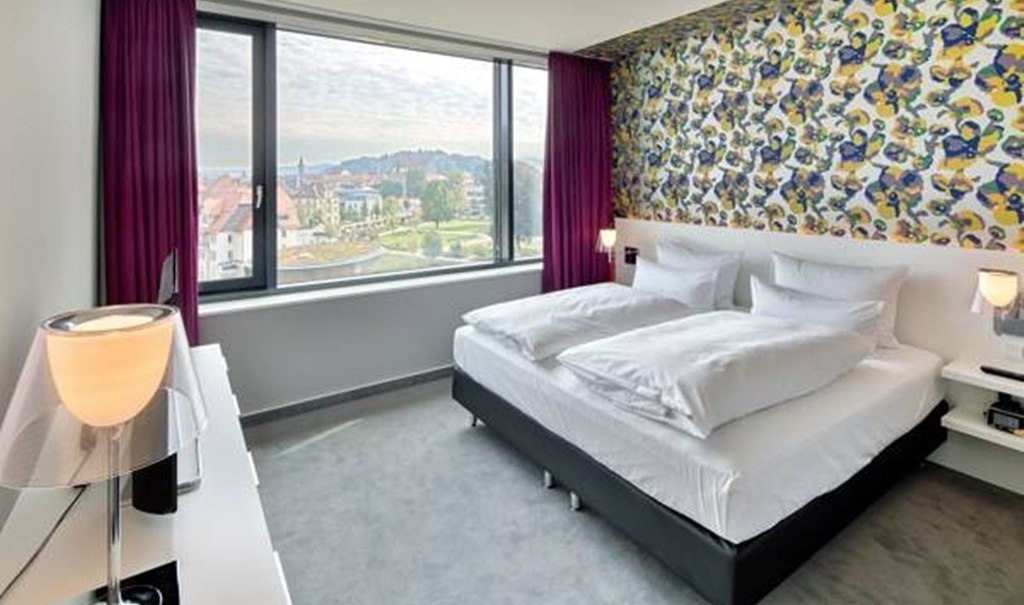 Modernes Hotelzimmer mit Doppelbett, bunter Tapete und großem Fenster mit Stadtblick. Lila Vorhänge und stilvolle Lampen ergänzen das Design., © Hotel am Remspark Modernes Hotelzimmer mit Doppelbett, bunter Tapete und großem Fenster mit Stadtblick. Lila Vorhänge und stilvolle Lampen ergänzen das Design., © Hotel am Remspark