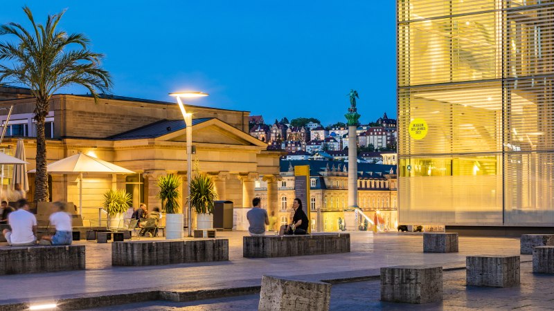 Abendstimmung am Kleinen Schlossplatz mit Palmen, Menschen und moderner Architektur. Im Hintergrund sind beleuchtete Gebäude und eine Statue zu sehen., © SMG, Werner Dieterich Abendstimmung am Kleinen Schlossplatz mit Palmen, Menschen und moderner Architektur. Im Hintergrund sind beleuchtete Gebäude und eine Statue zu sehen., © SMG, Werner Dieterich
