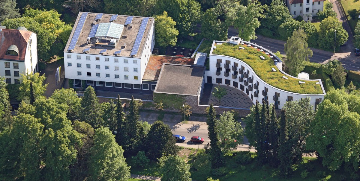Luftaufnahme eines modernen Gebäudekomplexes mit begrüntem Dach, umgeben von Bäumen und Grünflächen. Solarpaneele sind auf dem Dach sichtbar., © TOMAS