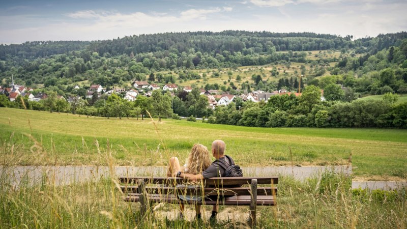 Ein Paar sitzt auf einer Bank und blickt auf eine grüne Landschaft mit einem Dorf im Hintergrund., © Natur.Nah. Schönbuch & Heckengäu