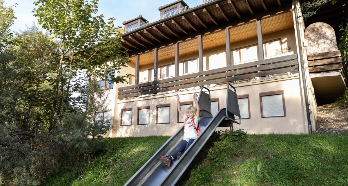 Ein Kind rutscht fröhlich eine Rutsche hinunter. Im Hintergrund steht ein großes Gebäude mit Holzbalkon und mehreren Fenstern., © Foto Thomas Zehnder Ein Kind rutscht fröhlich eine Rutsche hinunter. Im Hintergrund steht ein großes Gebäude mit Holzbalkon und mehreren Fenstern., © Foto Thomas Zehnder