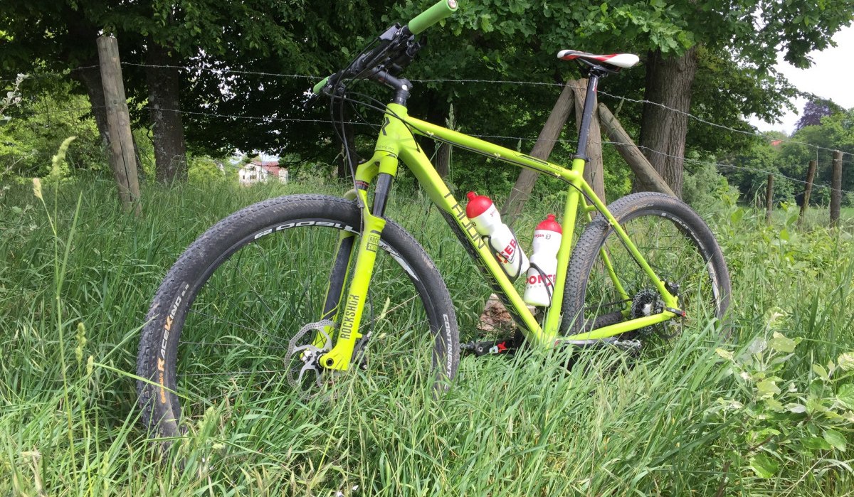 Grünes Mountainbike im hohen Gras neben einem Holzzaun, umgeben von Bäumen., © www.pro-cycl.de