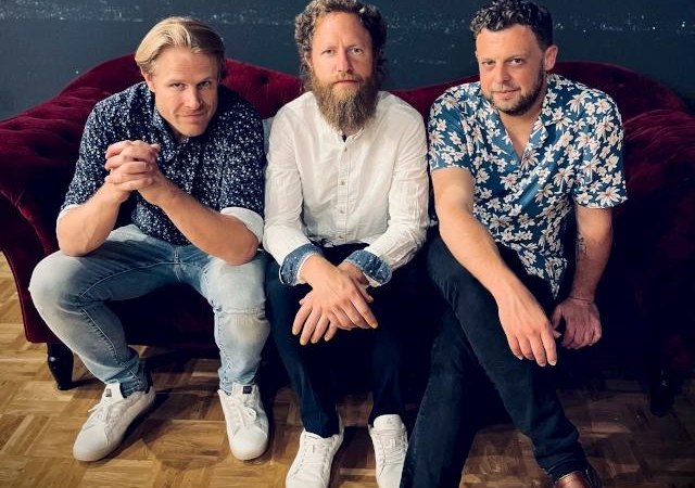 Drei Männer sitzen auf einem roten Sofa, lässig gekleidet in Hemden und Jeans, und blicken in die Kamera., © Foto Daniel Mudrack Drei Männer sitzen auf einem roten Sofa, lässig gekleidet in Hemden und Jeans, und blicken in die Kamera., © Foto Daniel Mudrack