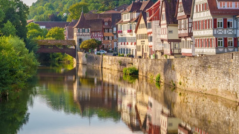 Fachwerkhäuser in Schwäbisch Hall spiegeln sich im Fluss. Eine Brücke und grüne Hügel im Hintergrund vervollständigen die malerische Szene., © Michael Kühneisen Fachwerkhäuser in Schwäbisch Hall spiegeln sich im Fluss. Eine Brücke und grüne Hügel im Hintergrund vervollständigen die malerische Szene., © Michael Kühneisen