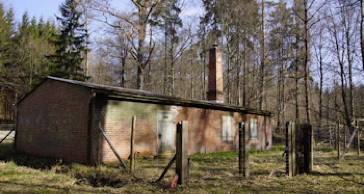 Ein altes Backsteingebäude mit Schornstein steht in einem lichten Wald. Ein verfallener Zaun umgibt das Gebäude. Ein altes Backsteingebäude mit Schornstein steht in einem lichten Wald. Ein verfallener Zaun umgibt das Gebäude.