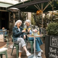 Zwei Frauen sitzen in einem gem&uuml;tlichen Caf&eacute; im Freien, sto&szlig;en mit Getr&auml;nken an und lachen. Ein Schild bietet Zimtschnecken an., &copy; Stuttgart Marketing GmbH - Sarah Schmid