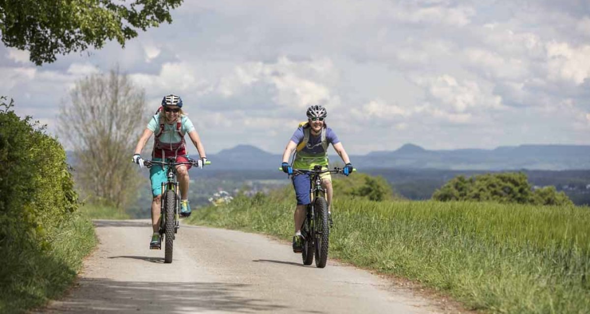 Zwei Radfahrer auf einem ländlichen Weg, umgeben von grünen Feldern und Hügeln im Hintergrund. Der Himmel ist bewölkt., © Schwäbische Alb Tourismusverband e.V. Zwei Radfahrer auf einem ländlichen Weg, umgeben von grünen Feldern und Hügeln im Hintergrund. Der Himmel ist bewölkt., © Schwäbische Alb Tourismusverband e.V.