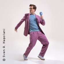 Eine Person in einem lila Anzug und blauen Hemd h&auml;lt ein Mikrofon und posiert dynamisch vor einem wei&szlig;en Hintergrund., &copy; links im Bild