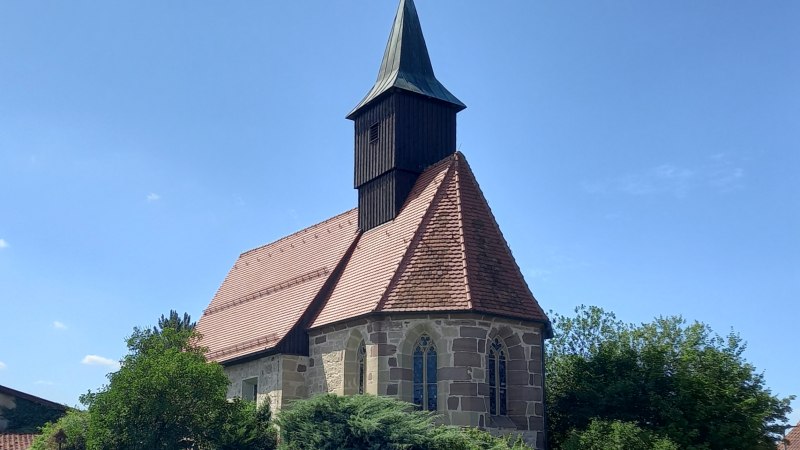 Kleine Kirche aus hellen Steinen mit dunkel verbrettertem Dachreiter auf rotbraunem Zigeldach, Blick auf den Chor mit gotischen Fenstern und die Südseite der Kirche, von Bäumen und Gebüsch umgeben, © Petra Natzkowski