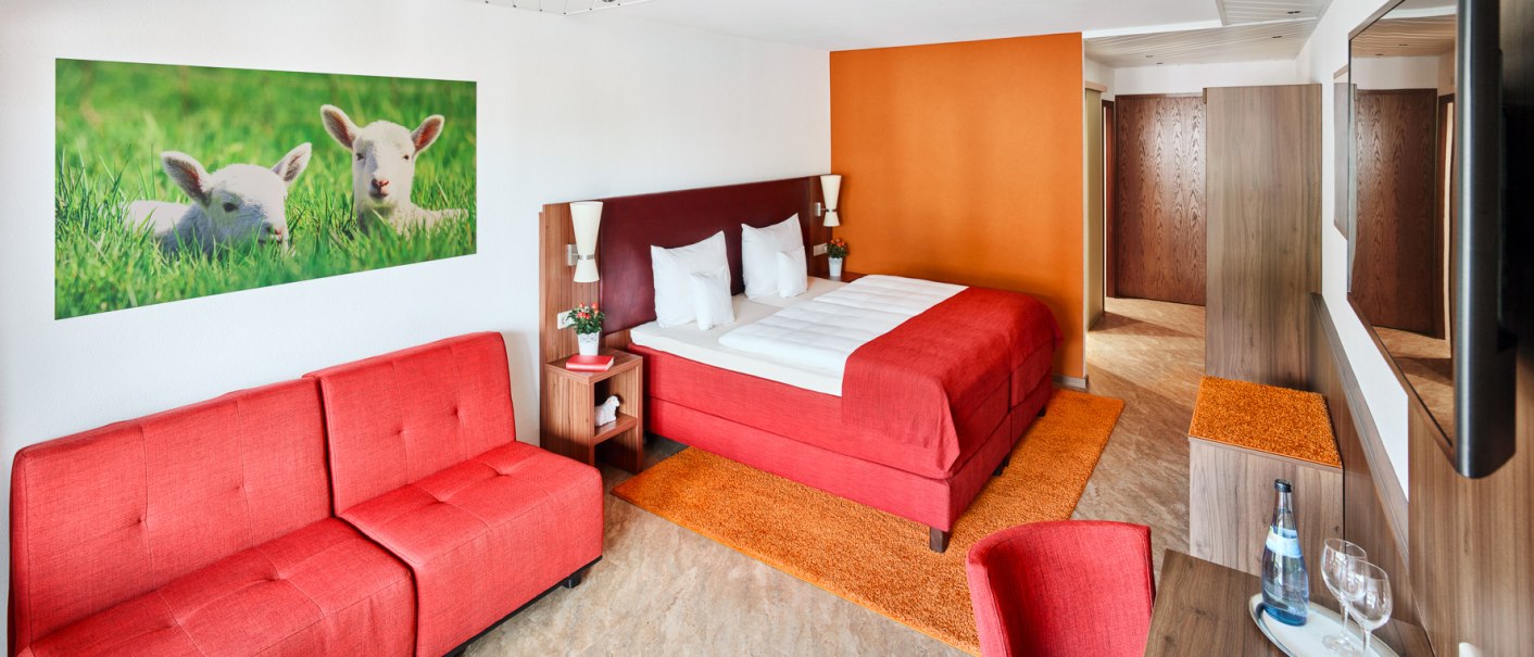 Modernes Hotelzimmer mit rotem Doppelbett, Sofa, Tisch und Wandbild von Lämmern. Helle, freundliche Einrichtung mit orangen Akzenten., © Hotel Restaurant Lamm