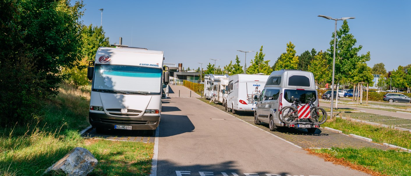 Wohnmobilstellplatz in Fellbach mit geparkten Wohnmobilen und Autos, umgeben von Bäumen und klarem Himmel. Auf der Straße steht 'Feuerwehrzufahrt'., © Stuttgart-Marketing GmbH, Thomas Niedermüller Wohnmobilstellplatz in Fellbach mit geparkten Wohnmobilen und Autos, umgeben von Bäumen und klarem Himmel. Auf der Straße steht 'Feuerwehrzufahrt'., © Stuttgart-Marketing GmbH, Thomas Niedermüller