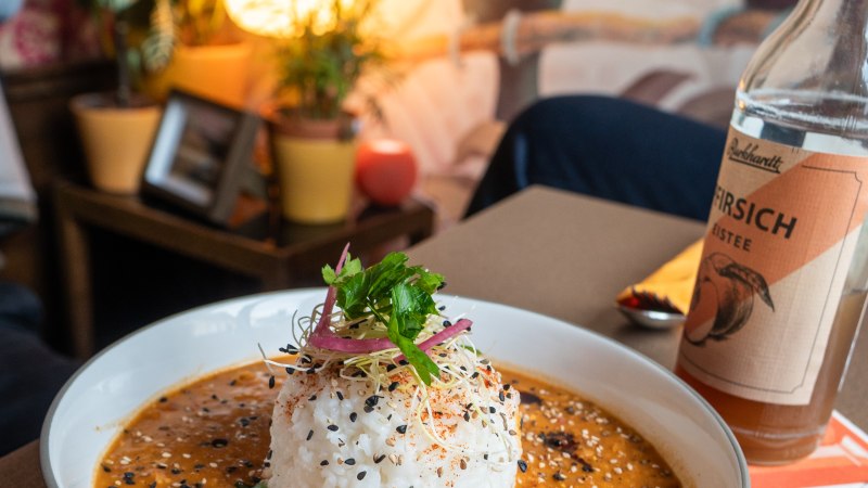 Ein Teller mit Reis und Curry, garniert mit Sesam und Kr&auml;utern, steht auf einem Tisch. Im Hintergrund sind Pflanzen und ein Wandbild mit Tukanen zu sehen., &copy; HerzoginN&uuml;rtingen