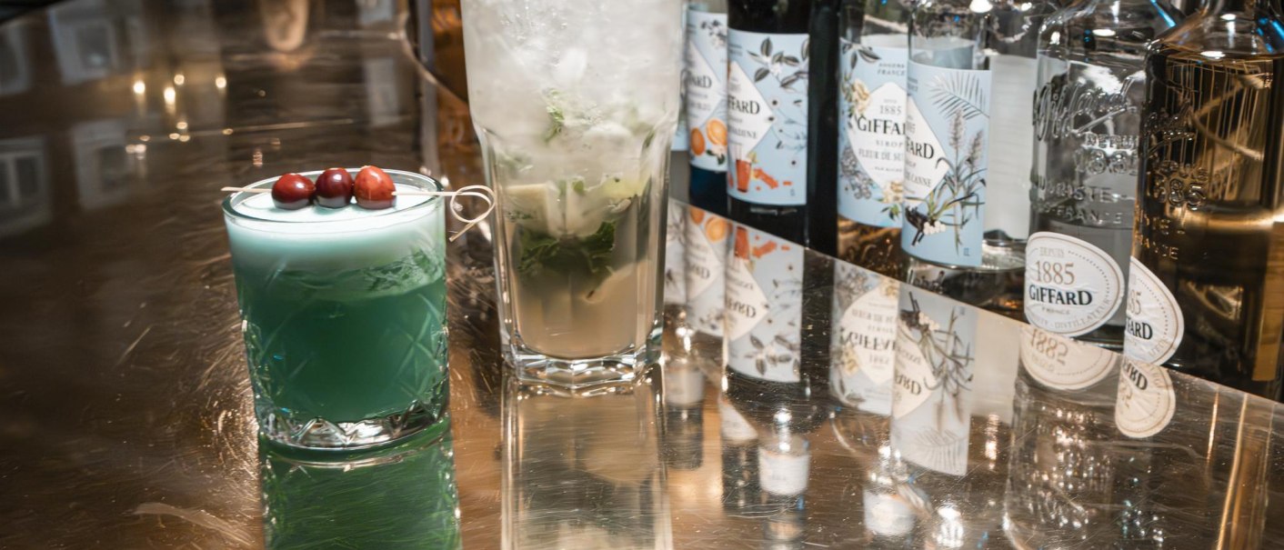 Zwei Cocktails auf einer glänzenden Bar. Links ein blauer Drink mit Beeren, rechts ein Mojito. Im Hintergrund stehen Flaschen mit Likören., © SMG Stuttgart Marketing GmbH - Sarah Schmid