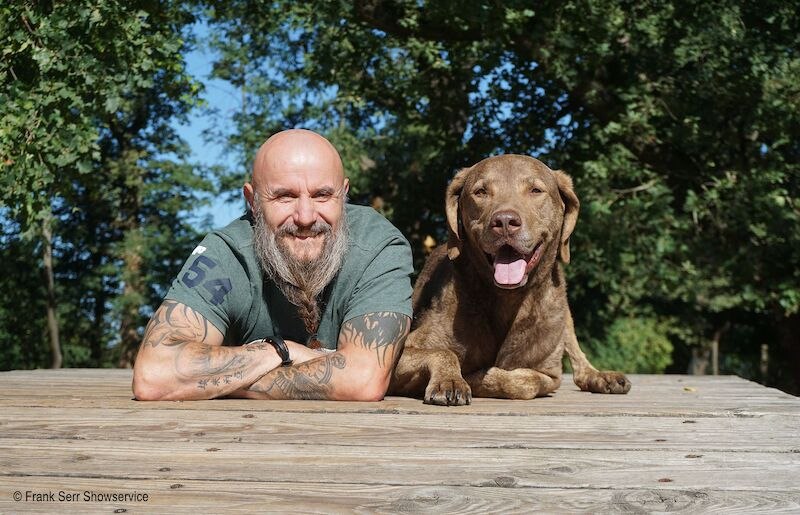 Ein Mann mit Bart und Tattoos liegt lächelnd neben einem braunen Hund auf einem Holztisch im Freien. Beide wirken entspannt und glücklich., © Stadthalle Leonberg Ein Mann mit Bart und Tattoos liegt lächelnd neben einem braunen Hund auf einem Holztisch im Freien. Beide wirken entspannt und glücklich., © Stadthalle Leonberg