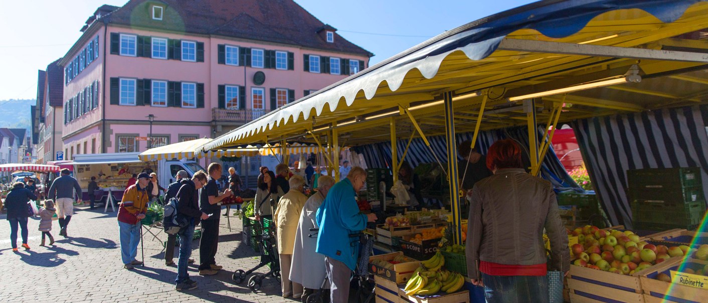 Menschen kaufen auf einem Markt in Murrhardt ein. Gelbe Marktstände bieten Obst und Gemüse an. Ein großes, rosa Gebäude im Hintergrund., © Stuttgart-Marketing GmbH