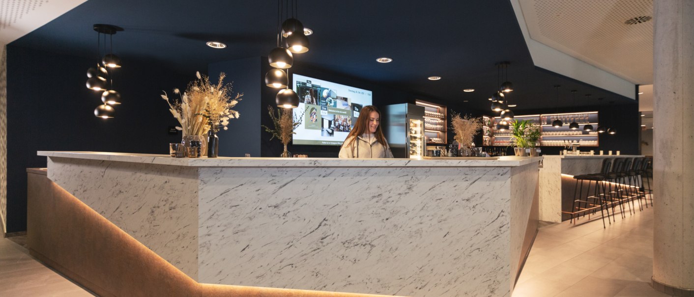 Moderne Rezeption mit Marmor-Theke, stilvoller Beleuchtung und Dekoration. Eine Person steht hinter der Theke, im Hintergrund ist eine Bar zu sehen., © the niu Kettle - Novum Hospitality
