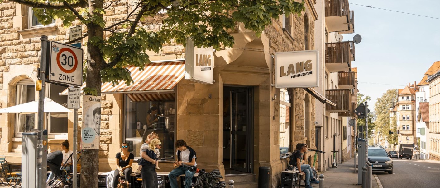 Café Lang an einer Straßenecke mit Menschen, die draußen sitzen. Ein 30er-Zone-Schild und Bäume sind ebenfalls zu sehen., © SMG, Sarah Schmid Café Lang an einer Straßenecke mit Menschen, die draußen sitzen. Ein 30er-Zone-Schild und Bäume sind ebenfalls zu sehen., © SMG, Sarah Schmid