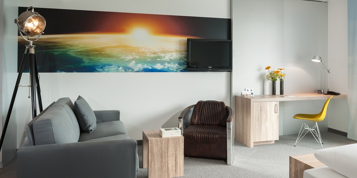 Modernes Hotelzimmer mit grauem Sofa, Ledersessel, Schreibtisch, gelbem Stuhl und großem Wandbild eines Sonnenaufgangs über der Erde., © Essential by Dorint