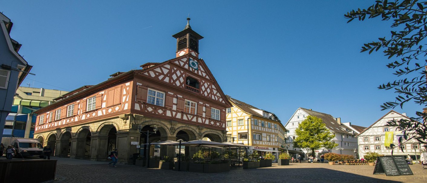 Fachwerkhäuser am Waiblinger Marktplatz bei sonnigem Wetter. Menschen flanieren, ein Café mit Sonnenschirmen ist zu sehen. Der Himmel ist klar und blau., © SMG, Sarah Schmid Fachwerkhäuser am Waiblinger Marktplatz bei sonnigem Wetter. Menschen flanieren, ein Café mit Sonnenschirmen ist zu sehen. Der Himmel ist klar und blau., © SMG, Sarah Schmid