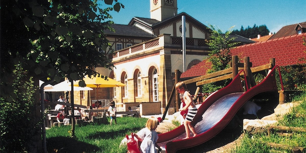 Kinder spielen auf einer Rutsche vor einem historischen Gebäude mit Turm und Sonnenschirmen im Garten., © Stuttgart-Marketing GmbH Kinder spielen auf einer Rutsche vor einem historischen Gebäude mit Turm und Sonnenschirmen im Garten., © Stuttgart-Marketing GmbH