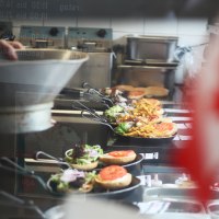 In einer Küche werden Burger und Pommes in Pfannen zubereitet. Im Vordergrund ist unscharf eine kanadische Flagge zu sehen., © SMG