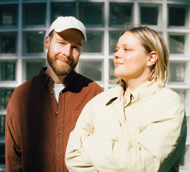 Zwei Personen vor einer Glaswand, eine trägt eine Mütze und ein braunes Hemd, die andere eine helle Jacke. Beide lächeln in die Sonne., © Scala Kultur Live gGmbH Zwei Personen vor einer Glaswand, eine trägt eine Mütze und ein braunes Hemd, die andere eine helle Jacke. Beide lächeln in die Sonne., © Scala Kultur Live gGmbH