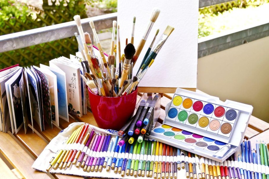 Ein Tisch mit Malutensilien: Pinsel in einem roten Becher, Buntstifte, Filzstifte, Wasserfarben und ein aufgeschlagenes Buch., © © Astrid Untermann