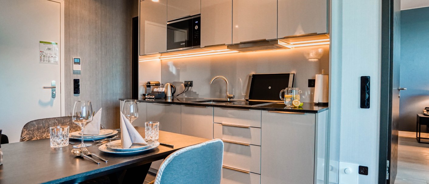 Moderne Küche in einer Suite mit Esstisch, gedeckt für zwei Personen. Helle Schränke, Beleuchtung unter den Schränken, Wasserkocher und Kaffeemaschine vorhanden., © CLOUD N°7 APARTMENTS GmbH