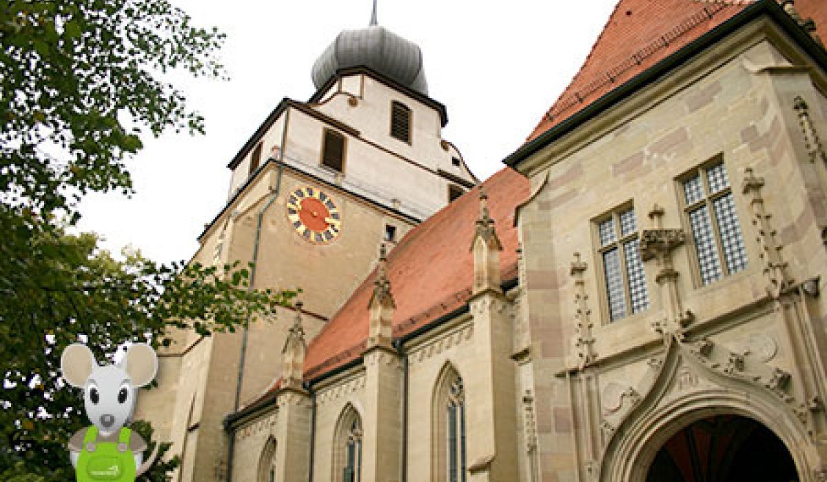 Eine Kirche mit einem markanten Uhrturm und einer Mausfigur im Vordergrund. Die Architektur ist gotisch gepr&auml;gt.