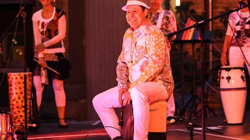 Ein Musiker sitzt lächelnd auf einer Cajon auf der Bühne, umgeben von anderen Musikern mit Congas. Die Szene ist in warmem Licht gehalten., © © PeterPöschl Ein Musiker sitzt lächelnd auf einer Cajon auf der Bühne, umgeben von anderen Musikern mit Congas. Die Szene ist in warmem Licht gehalten., © © PeterPöschl