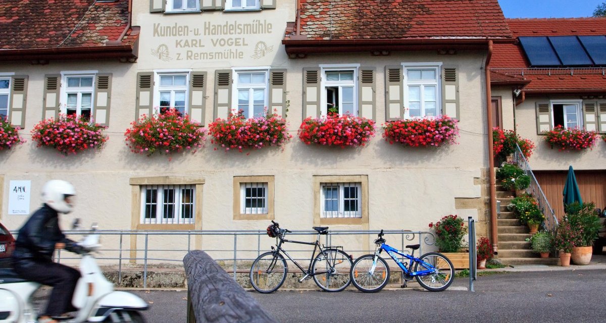 Ein traditionelles Gebäude mit roten Dachziegeln und Blumenkästen. Ein Rollerfahrer fährt vorbei, zwei Fahrräder stehen vor dem Haus., © Waiblingen - Stuttgart-Marketing GmbH Ein traditionelles Gebäude mit roten Dachziegeln und Blumenkästen. Ein Rollerfahrer fährt vorbei, zwei Fahrräder stehen vor dem Haus., © Waiblingen - Stuttgart-Marketing GmbH