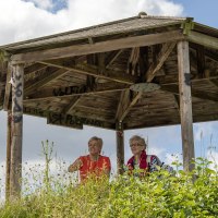 Zwei Personen sitzen in einem hölzernen Pavillon auf einem Hügel, umgeben von Pflanzen und bewölktem Himmel., © SMG, Achim Mende