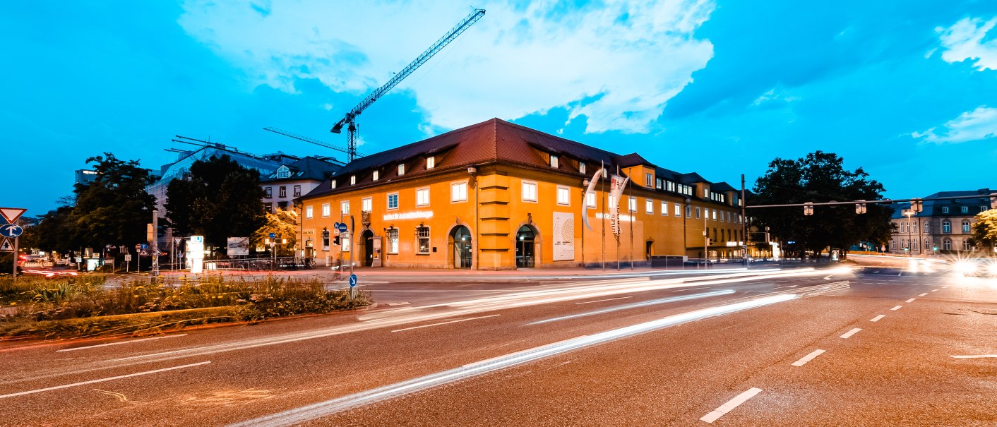 Gelbes Gebäude bei Abenddämmerung, beleuchtet von Straßenlichtern. Autos ziehen Lichtspuren auf der Straße, Baukräne im Hintergrund., © Luca Siermann, ifa Galerie Stuttgart