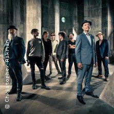 Neun Personen posieren in einem industriellen Raum, einige tragen Hüte und Anzüge. Die Atmosphäre ist stilvoll und modern., © links im Bild Neun Personen posieren in einem industriellen Raum, einige tragen Hüte und Anzüge. Die Atmosphäre ist stilvoll und modern., © links im Bild