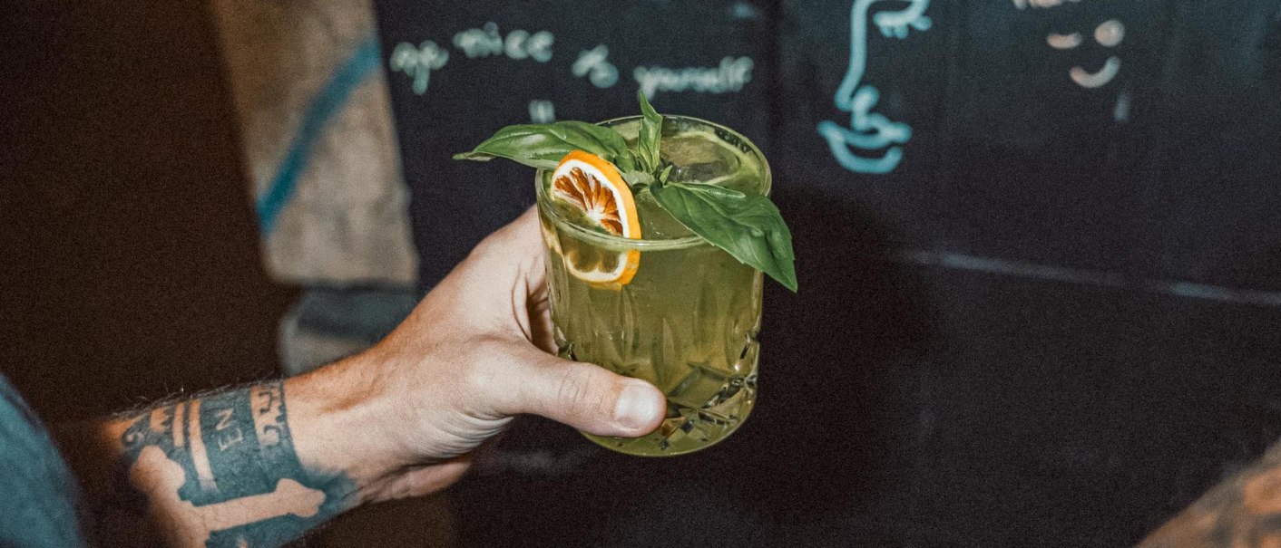 Eine tätowierte Hand hält ein grünes Getränk mit Basilikum und Orangenscheibe. Im Hintergrund ist eine Tafel mit Zeichnungen und Schrift zu sehen., © Bruno, Stuttgart
